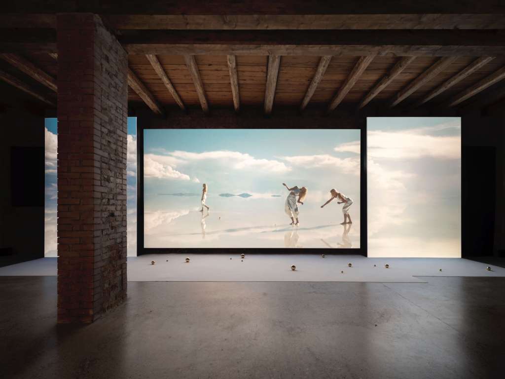 The image shows a dark room with three screens at the back wall and little balls lying in front of it. It is an image from the exhibition Lita Albuquerque.Liquid Light presented by bardoLa in Venice, 2022.