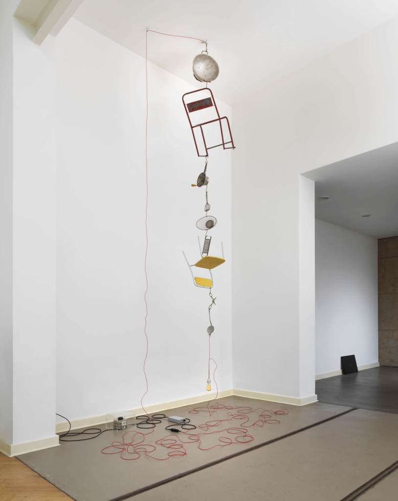 The image shows a mobile-like installation hanging from the ceiling; it is composed of chairs and various kitchen tools such as a grater, siege, and ladle. Under the structure, various cables are loosely lying around. The installation is a work by the artist Mona Hatoum.