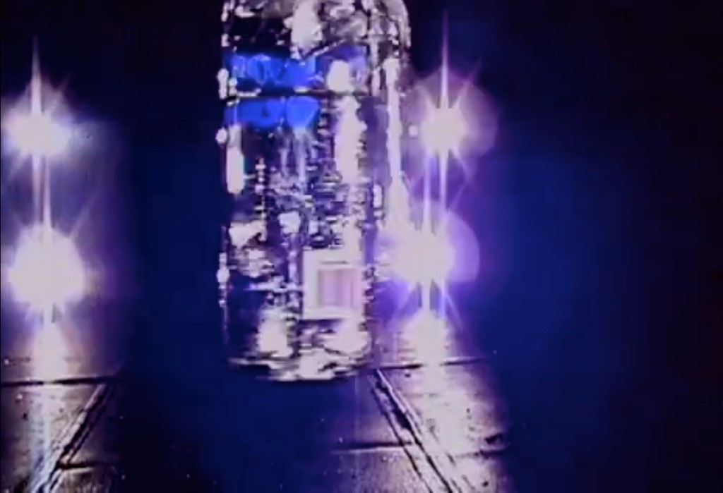 The image shows a glass bottle hanging from the top of the picture. The bottle is placed in the central front of the image, in the back five light sources can be seen. The whole image is blurry and in blue-black shades. It is a film still from the video "Walk With Me" by Rosemarie Trockel.