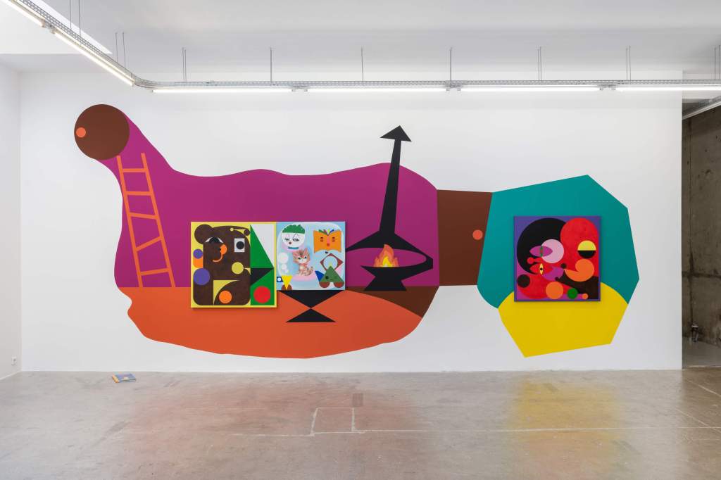 The image shows an open room with a white back wall on which a colorful mural is painted. The mural consists of various loosely geometric shapes forming a kind of rhythm. The image can be remotely interpreted as a housing scene. In the centre of the general shapes, three objects with smaller images are definable. It is an installation view of Ad Minoliti's exhibition "warm hole & hot tea" at Galerie Crèvecœur, 2023.