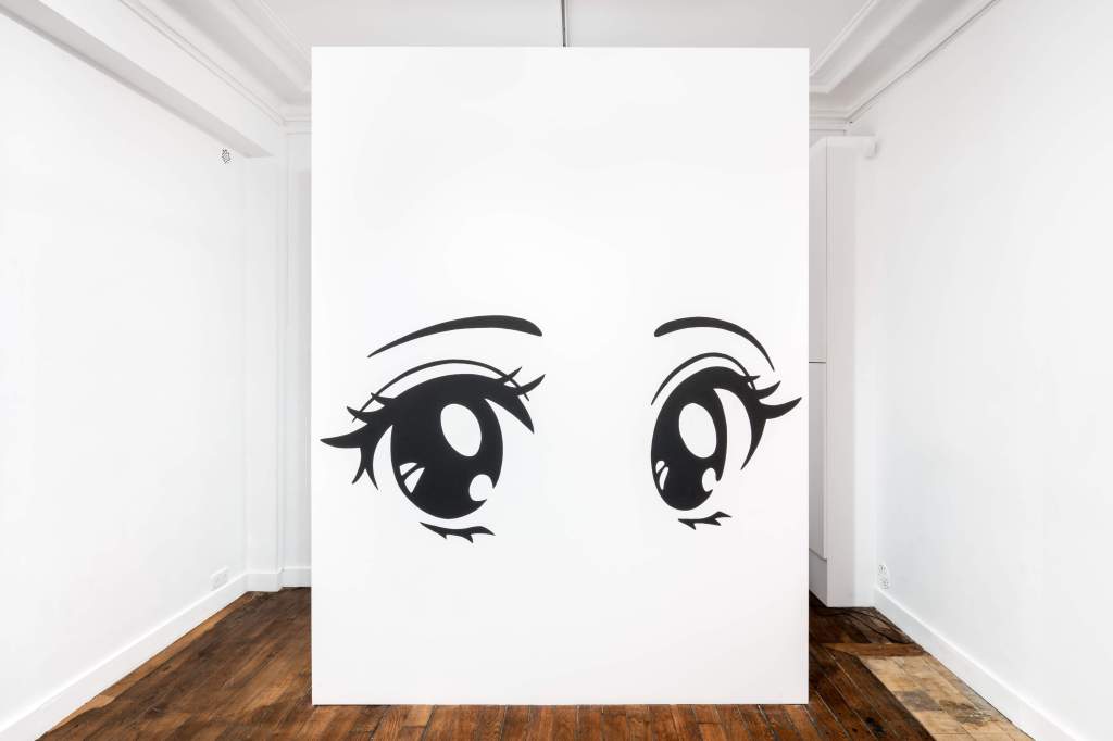 The image shows the fragment of a white wall standing in the centre of a room. Two black eyes are painted on the wall fragment. It is an installation view of Ad Minoliti's exhibition "warm hole & hot tea" at Galerie Crèvecœur, 2023.